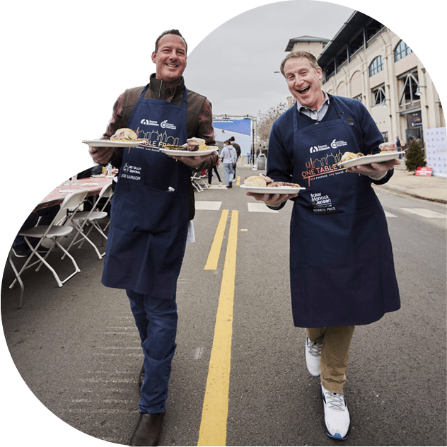 Two smiling men wearing blue aprons carry plates of food while walking outside on a street, with empty tables and chairs set up alongside a large building in the background.